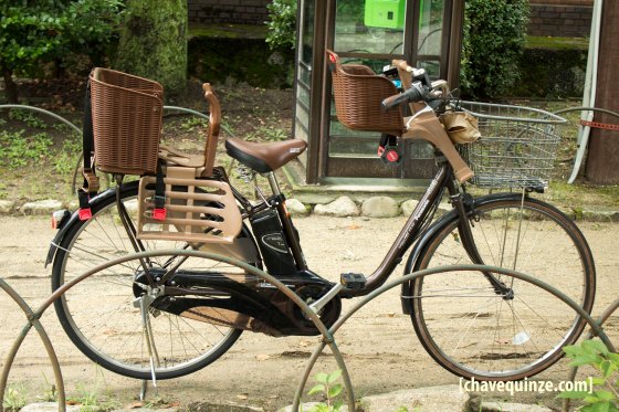 Bicicleta elétrica com duas cadeiras para criança e cesto, em frente à entrada do Castelo de Osaka.